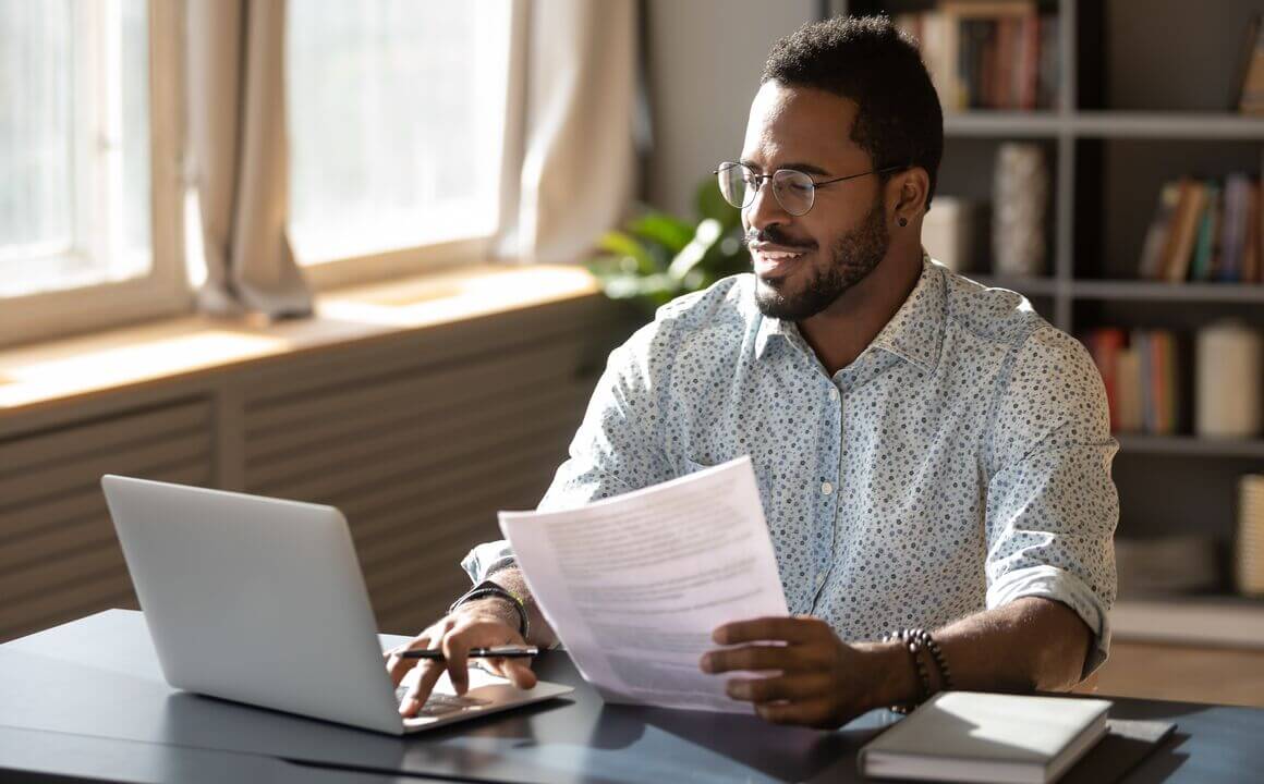 A smiling man reviews paperwork while typing at his laptop